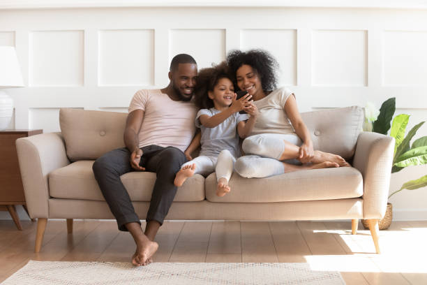 Smiling little mixed race girl holding smartphone, playing game, showing funny video to loving parents. Happy african american family sitting on comfortable couch in modern living room, using phone.